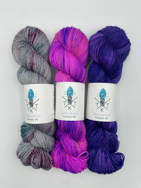 Three skeins of yarn in purple, pink, and gray with labels on a white background