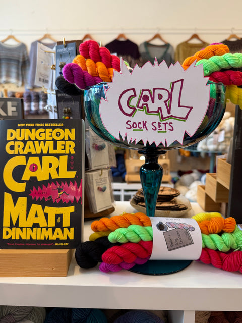A display of colorful yarn skeins and a sign reading 'CARL SOCK SETS' on a table in a retail or craft shop setting, with a book titled 'Dungeon Crawler Carl' by Matt Dinniman visible on the left.