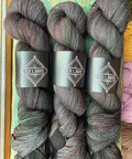 Three skeins of Ex Libris variegated yarn stacked vertically, featuring a dark charcoal gray and black base with subtle hints of olive green and muted burgundy. Each skein is wrapped in a black paper band displaying the Ex Libris logo.