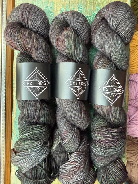 Three skeins of Ex Libris variegated yarn stacked vertically, featuring a dark charcoal gray and black base with subtle hints of olive green and muted burgundy. Each skein is wrapped in a black paper band displaying the Ex Libris logo.