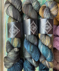 Six skeins of variegated yarn from Ex Libris, arranged in two rows on a rustic wooden surface. The yarn features variegated colors of slate blue, charcoal gray, black, and hints of olive green and gold.