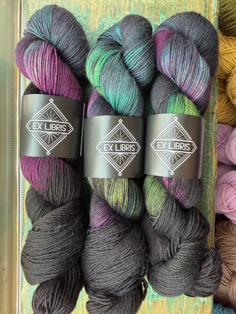 Three skeins of variegated yarn with dark gray, purple, and green color-shifting yarn, each wrapped with a black paper band labeled 'EX LIBRS'.