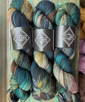 Three skeins of Ex Libris yarn stacked on a rustic wooden surface. The yarn features a variegated color pattern of deep teal, blue, brown, and hints of gold.
