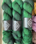 Three hanks of dark green and black variegated yarn with black paper bands labeled 'EX LIBRIS' arranged side by side on a rustic wooden surface.