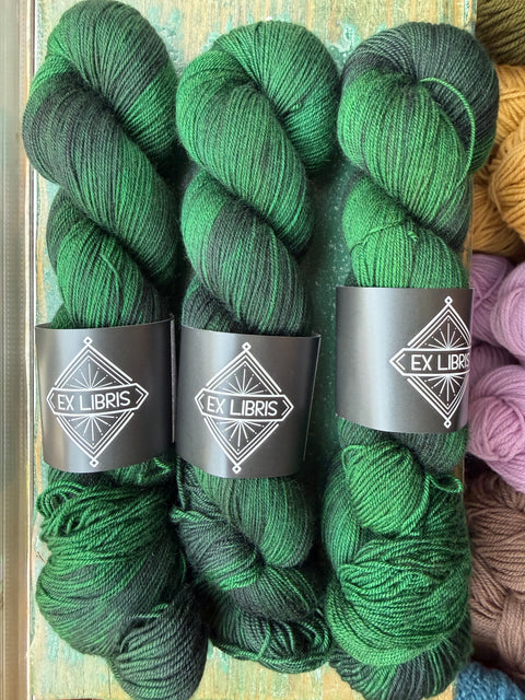 Three hanks of dark green and black variegated yarn with black paper bands labeled 'EX LIBRIS' arranged side by side on a rustic wooden surface.